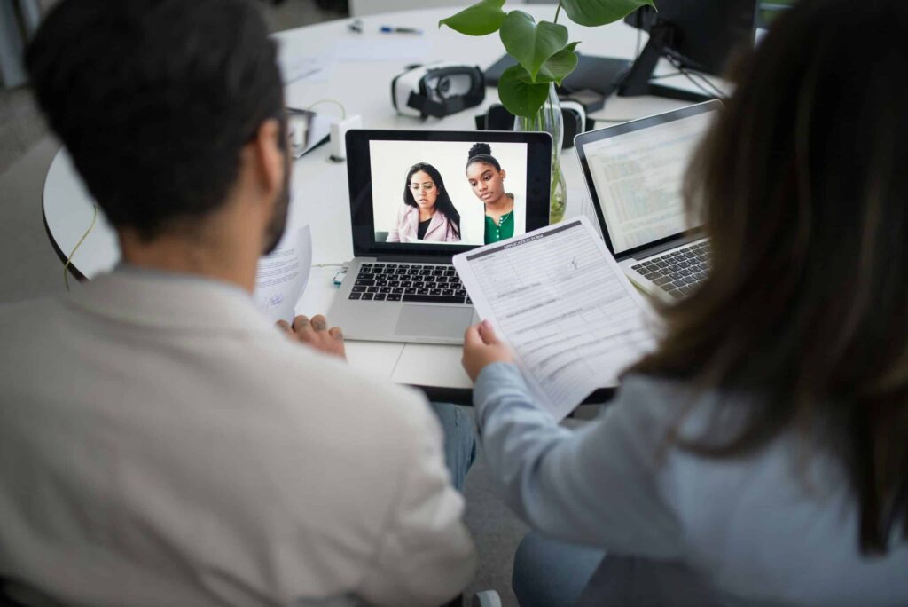 Two individuals are in a video call on a laptop, while another person holds a document at a modern workspace with plants.