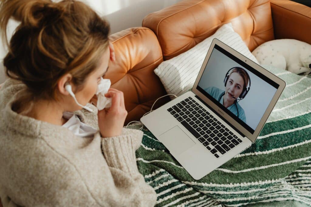 A person sits on a couch covered with a blanket, engaged in a video call on a laptop, wearing earphones.