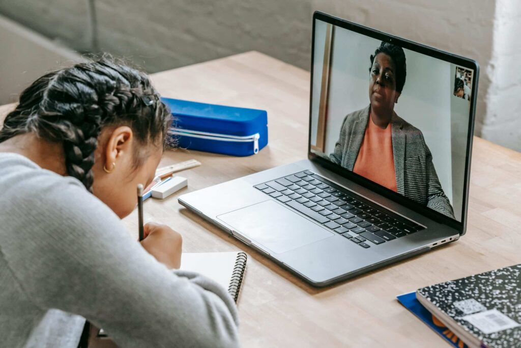 A student sits at a desk, focused on a laptop, engaging in a video call. School supplies and notebooks are on the table.