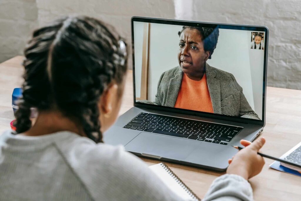 A young person with braids attentively engaging in a video call. The laptop screen shows a woman speaking, conveying a serious discussion.