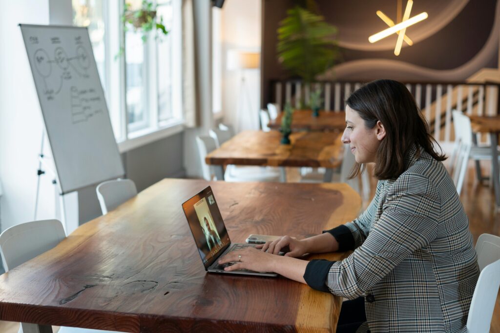 Woman in a checked blazer video calls on a laptop in a cozy office with wooden tables. A whiteboard and modern lighting add a professional touch.