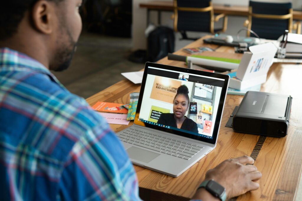 A man in a plaid shirt participates in a video call on a laptop at a wooden table. The laptop screen shows a woman speaking. The setting is a casual office space.