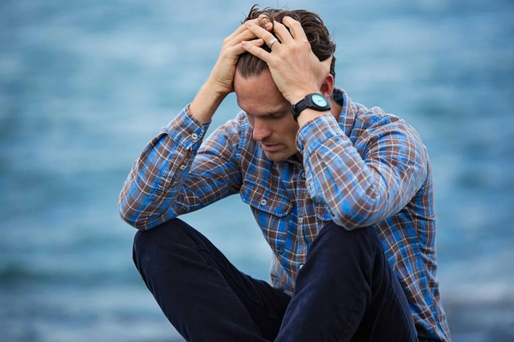 Man in plaid shirt sitting by the ocean, head in hands, appears stressed. Background is blurred blue water, conveying a sense of isolation or contemplation.