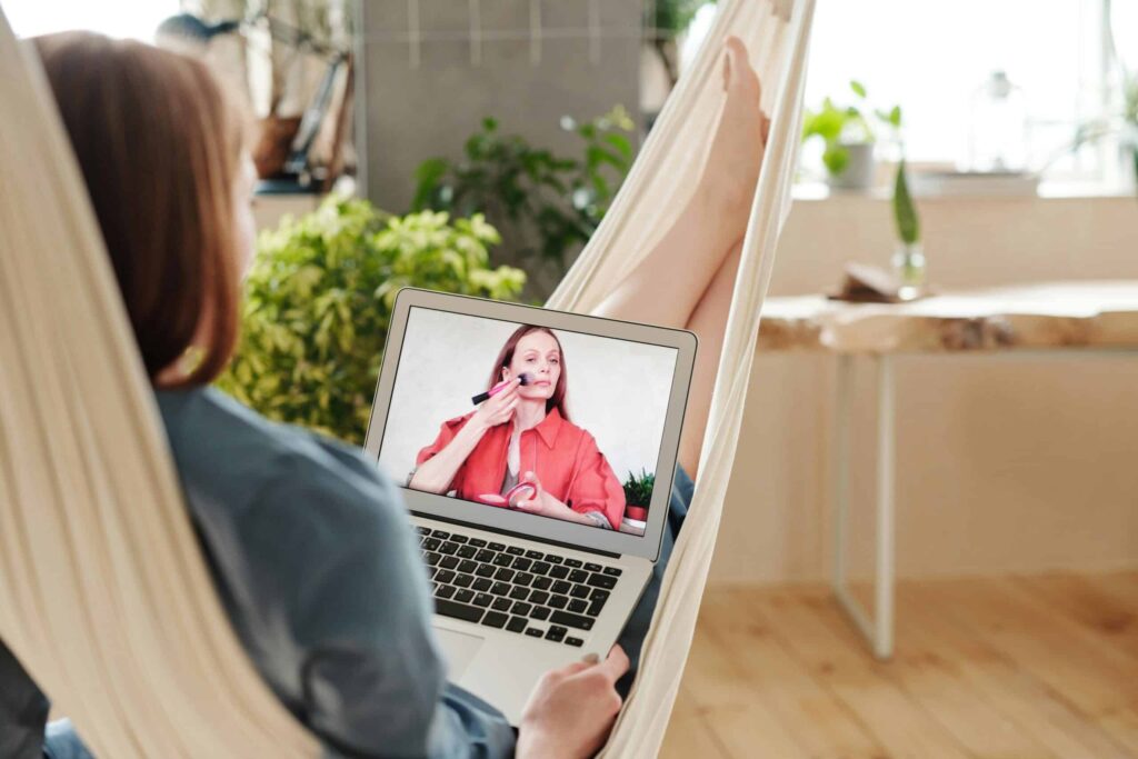 A person in a hammock watches a makeup tutorial on a laptop. The atmosphere is relaxed, with plants and soft lighting in a cozy room.