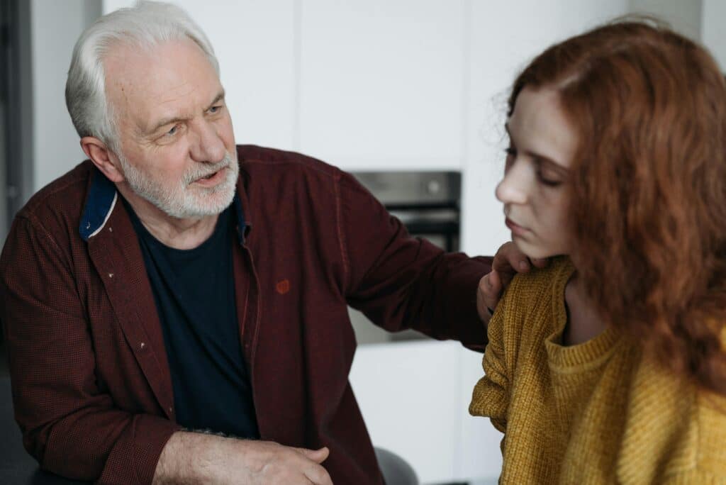 An older man with white hair and a beard comforts a young woman with red hair. He gently places a hand on her shoulder, conveying care and empathy.