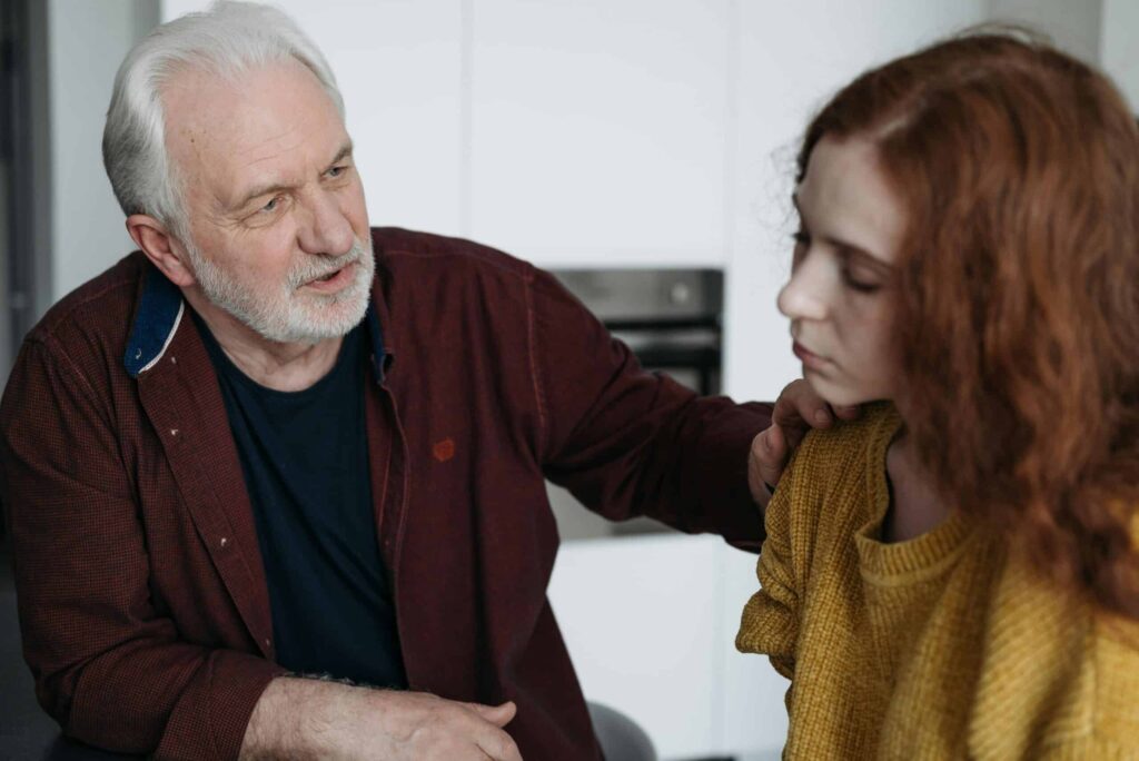 An older man with white hair and a beard comforts a young woman with red hair. He gently places a hand on her shoulder, conveying care and empathy.