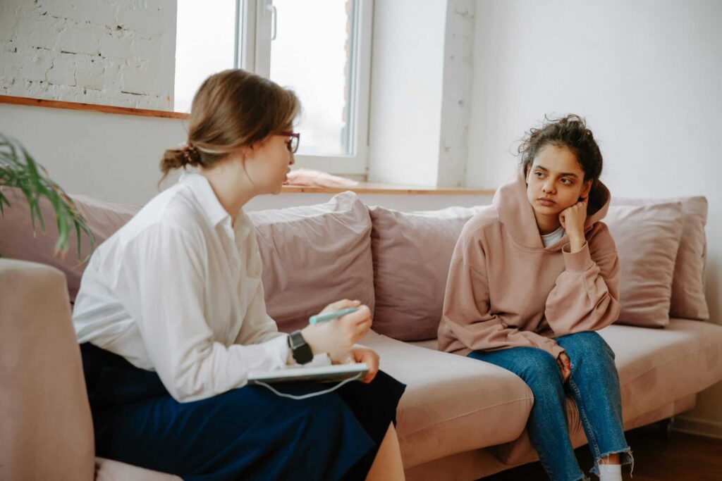 A woman in a white blouse holds a notebook while seated across from another woman in a pink hoodie, engaged in a conversation on a sofa.