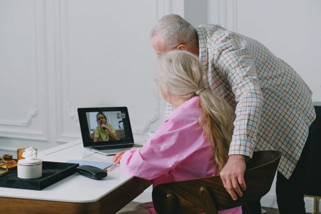 Elderly couple video calls a young woman on a laptop. The woman in pink sits while the man leans in, showing engagement and connection. Cozy, warm setting.