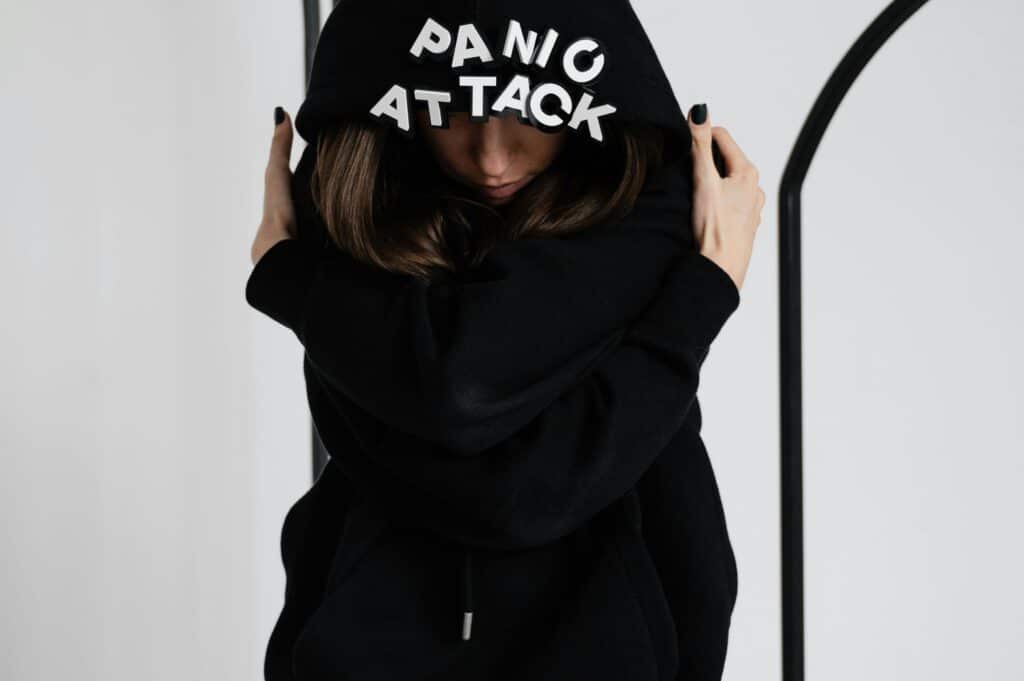 A person wearing a black hoodie with "Panic" and "Attack" on the back, embracing themselves against a simple background.
