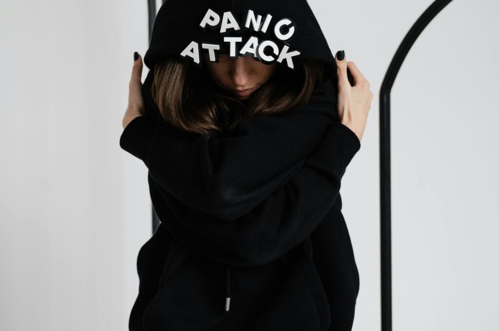 A person wearing a black hoodie with "Panic" and "Attack" on the back, embracing themselves against a simple background.
