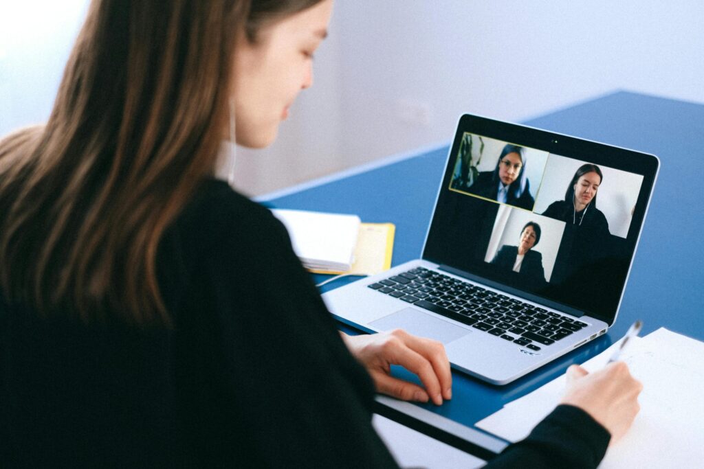 A person with long hair participates in a video conference on a laptop, taking notes and engaging with multiple remote participants.