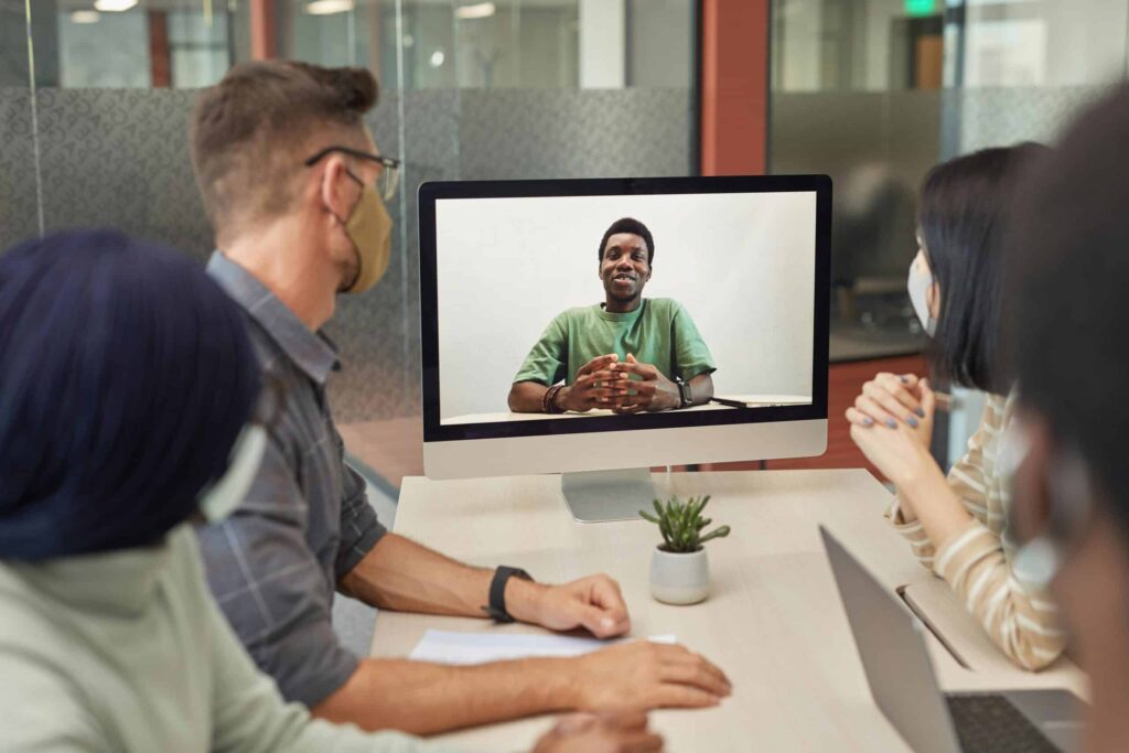 Four people in masks sitting around a table, engaged in a video call. On the screen, a person in a green shirt is speaking, conveying a collaborative tone.