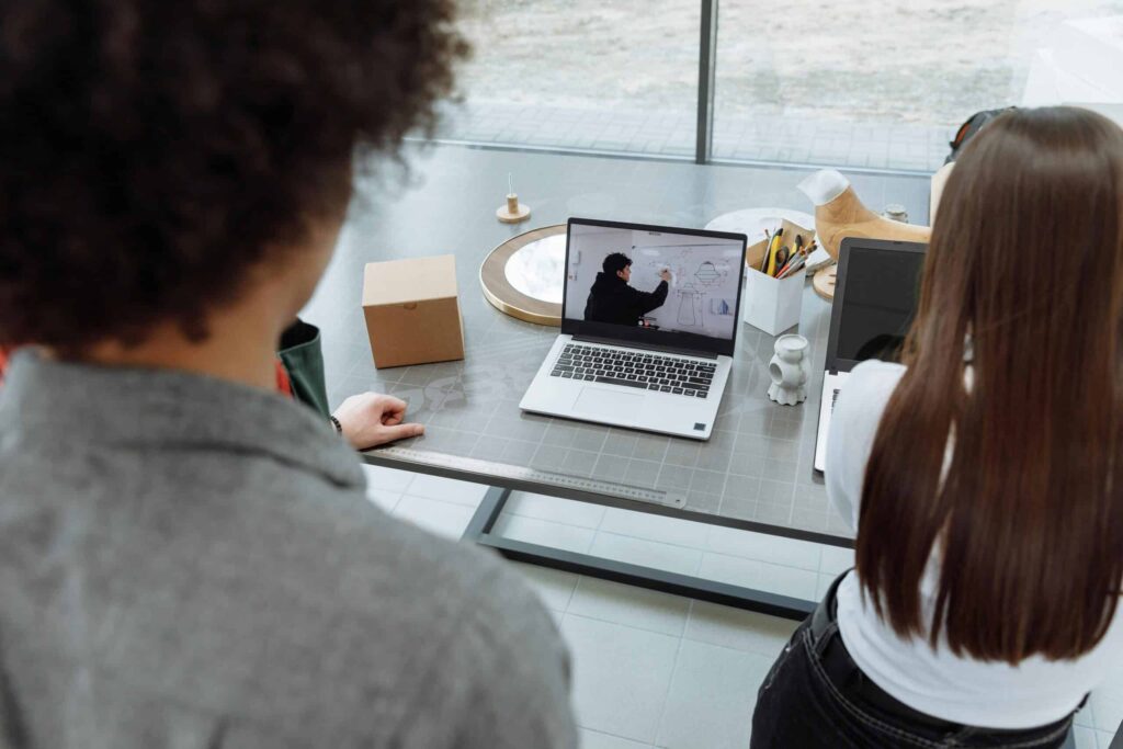 Two people stand by a table, watching a laptop screen showing a person writing on a whiteboard. The scene is calm and focused.