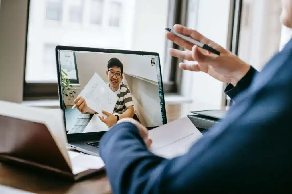 A person in a striped shirt holds documents during a video call, while another individual gestures with a pen, discussing remotely.