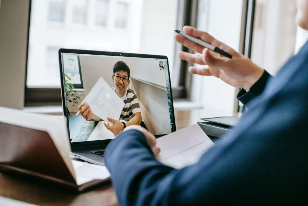 A person in a striped shirt holds documents during a video call, while another individual gestures with a pen, discussing remotely.