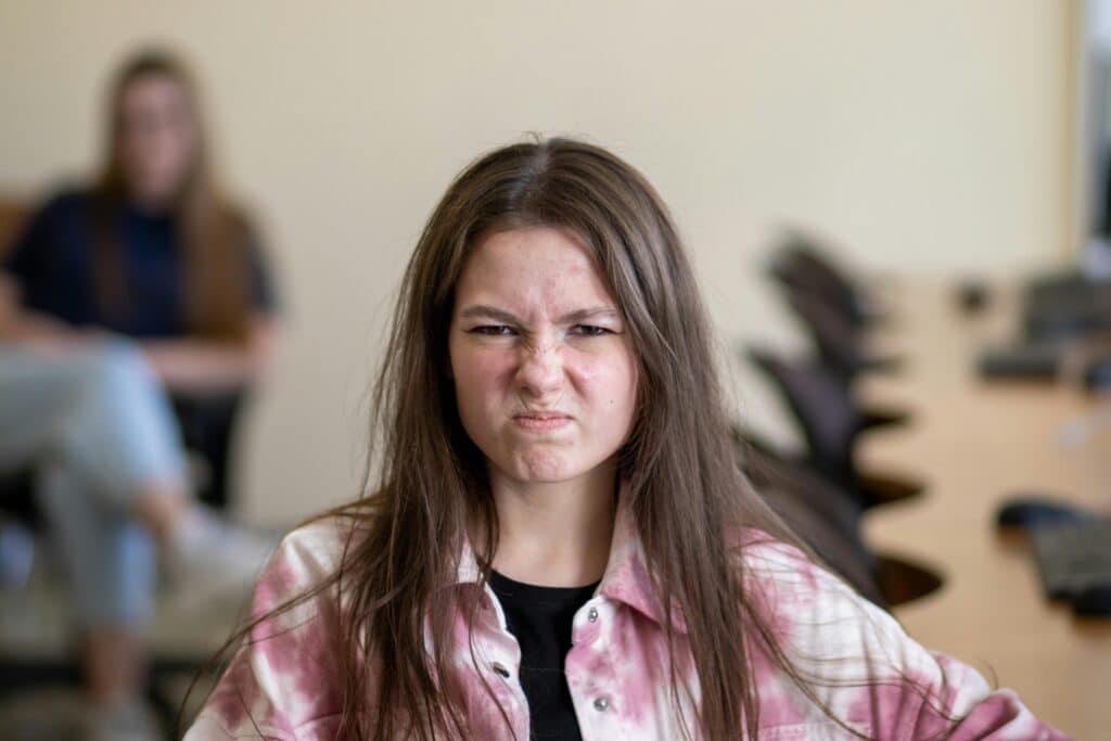 A young person with long hair wears a pink tie-dye shirt, seated in a classroom with another person in the background.