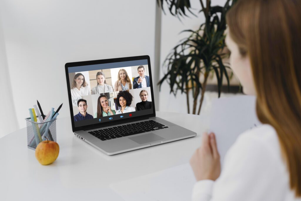 A woman participates in a video call on a laptop with eight people visible on the screen. An apple and pen holder sit on the table, creating a focused work atmosphere.