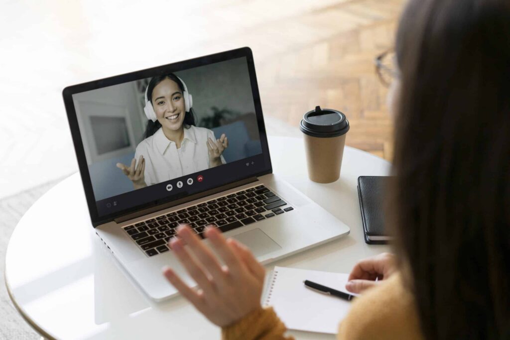 A person in a yellow sweater waves at a laptop screen during a video call. A smiling person with headphones is on the screen. A notepad and coffee cup are nearby.