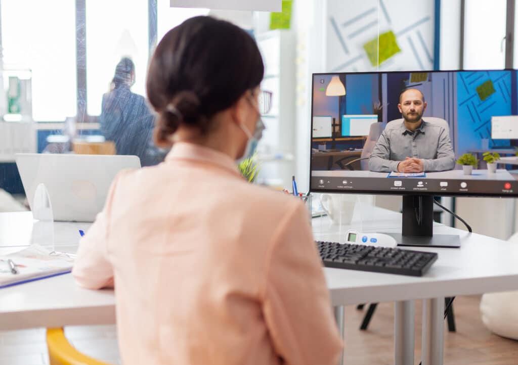 A woman in a mask participates in a video conference at her desk, viewing a man on a monitor in a modern, bright office setting.