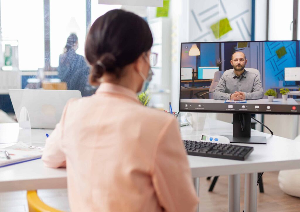 A woman in a mask participates in a video conference at her desk, viewing a man on a monitor in a modern, bright office setting.
