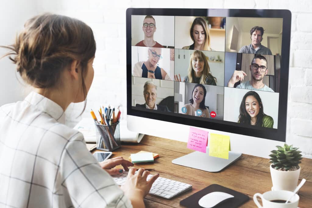 A woman participates in a virtual meeting on a desktop computer, displaying nine diverse, smiling people in a video call. She types notes, exuding focus and engagement.