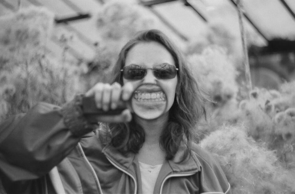 3:28 PMWoman in sunglasses grinning while fist-bumping someone in a greenhouse, black and white photo.