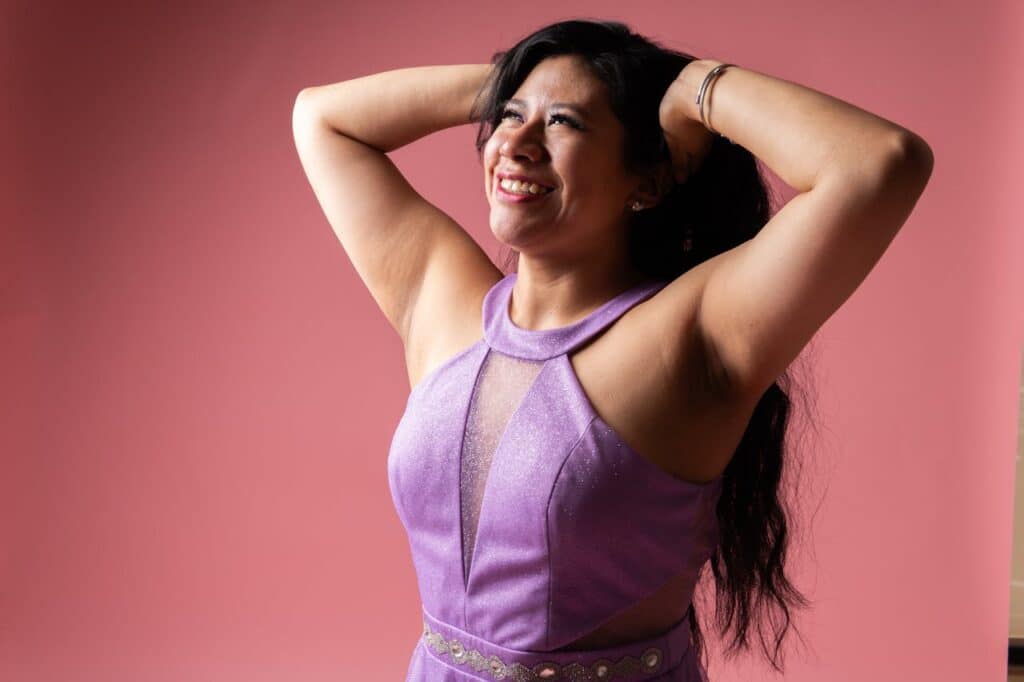 Smiling woman in a sparkly purple halter dress with arms raised behind her head against a pink background