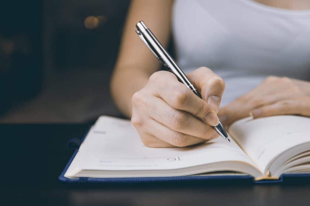 A person in a white tank top writes in an open journal with a silver pen, capturing the reflective practice of therapeutic journaling.