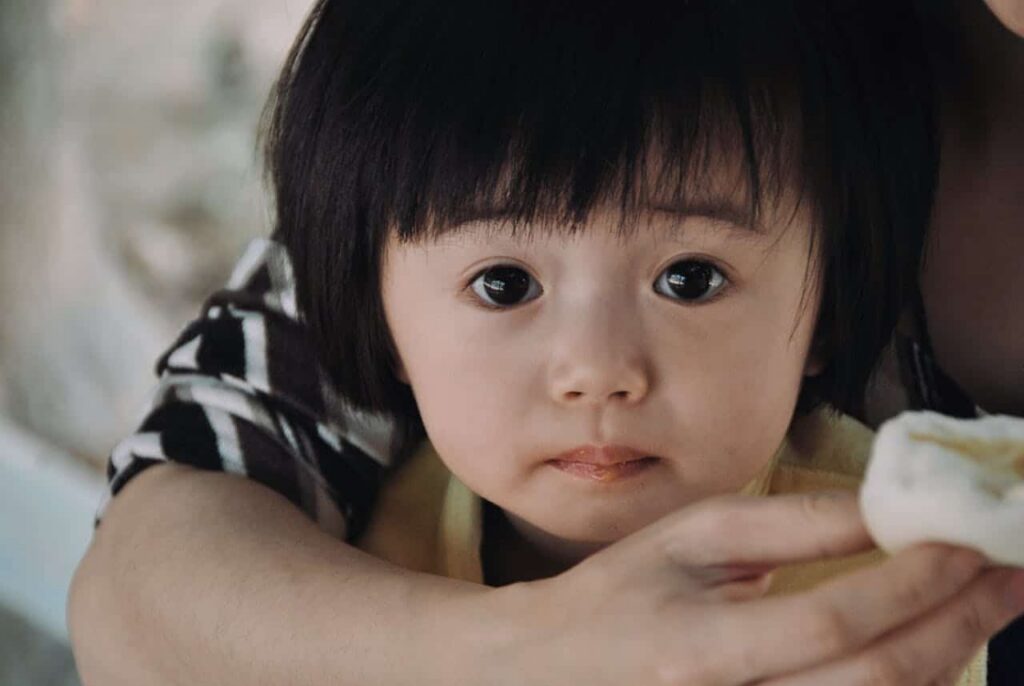 Toddler with dark hair held by an adult's arms, looking at the camera with a gentle expression.