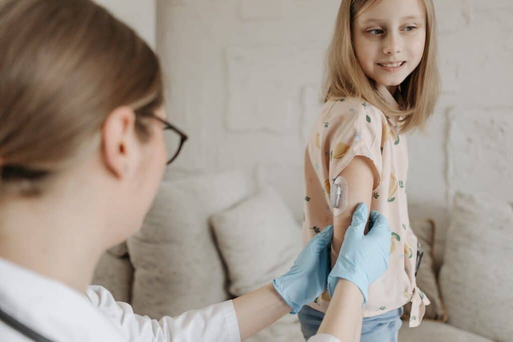A young girl smiles as a healthcare provider in blue gloves checks a glucose monitor on her arm.
