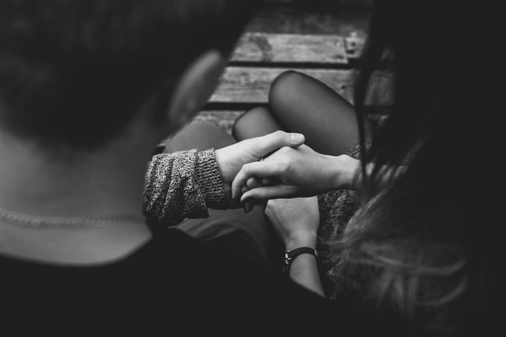 A black and white close-up of two people holding hands across a wooden table, conveying comfort and emotional support.