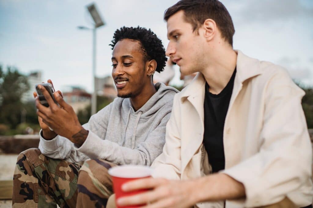 Two friends sitting outside, one showing something on his phone to the other while they share a moment together.