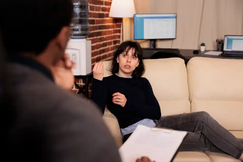 A woman on a beige couch gestures while talking to an unseen person holding a clipboard. A calm discussion environment with desks and monitors is in the background.